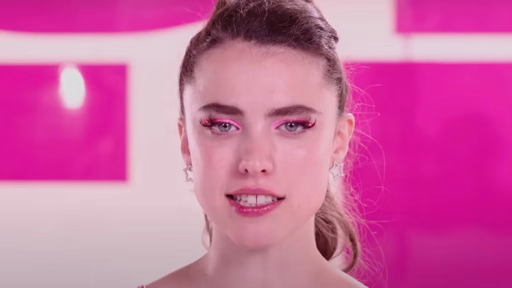 Margaret Qualley faces the camera against a bright pink background in The Substance, wearing dramatic makeup and earrings.