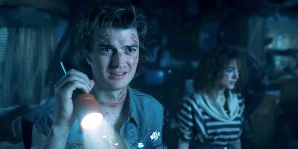 Steve holds a flashlight while Nancy stands behind him in a dark blue-tinted scene from Stranger Things Season 4.