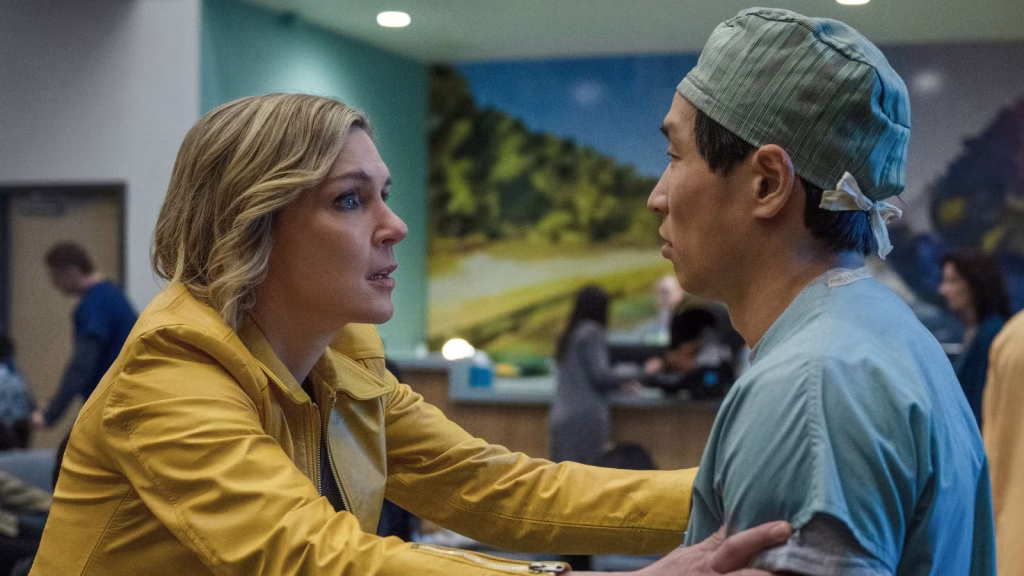 A woman in a yellow jacket grips a man in scrubs by the arms while speaking to him in a hospital setting in Pluribus.