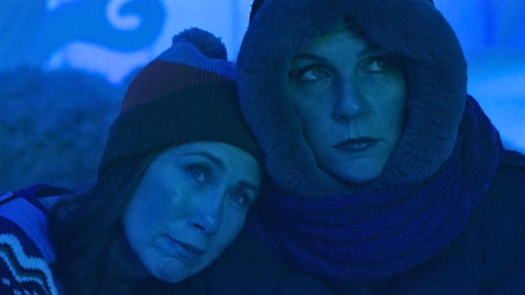 Two women in winter clothing lean close together in a blue-lit scene from Pluribus.