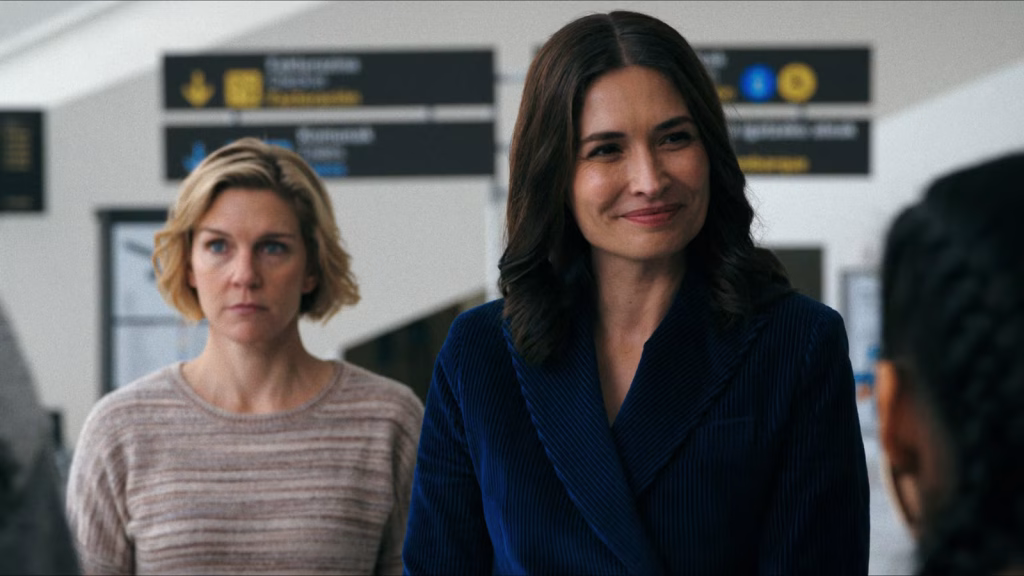 Two women stand indoors facing someone off camera, with one smiling calmly and the other watching more seriously in a scene from Pluribus.
