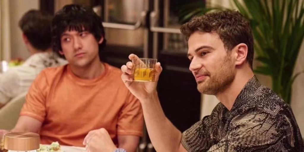Ethan and Cameron sit at a dining table indoors, with Cameron holding up a drink while Ethan looks on with a guarded expression.