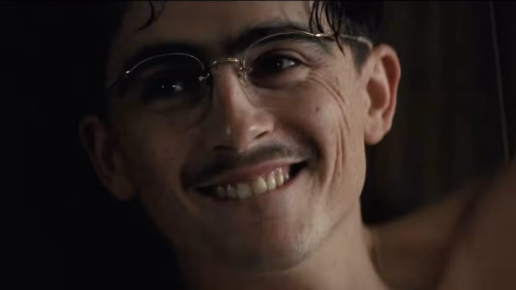 Close-up of a young man wearing wire-rim glasses and a thin mustache smiling in a dimly lit room.