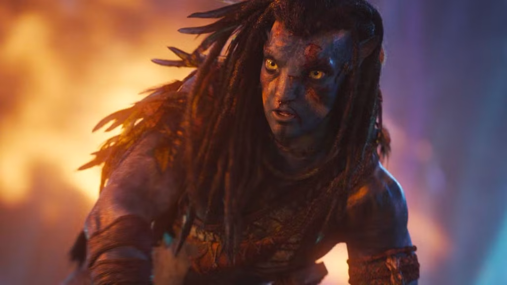 Jake Sully, a blue Na’vi man with braided hair, crouches and looks ahead in warm firelight with flames and smoke blurred in the background.