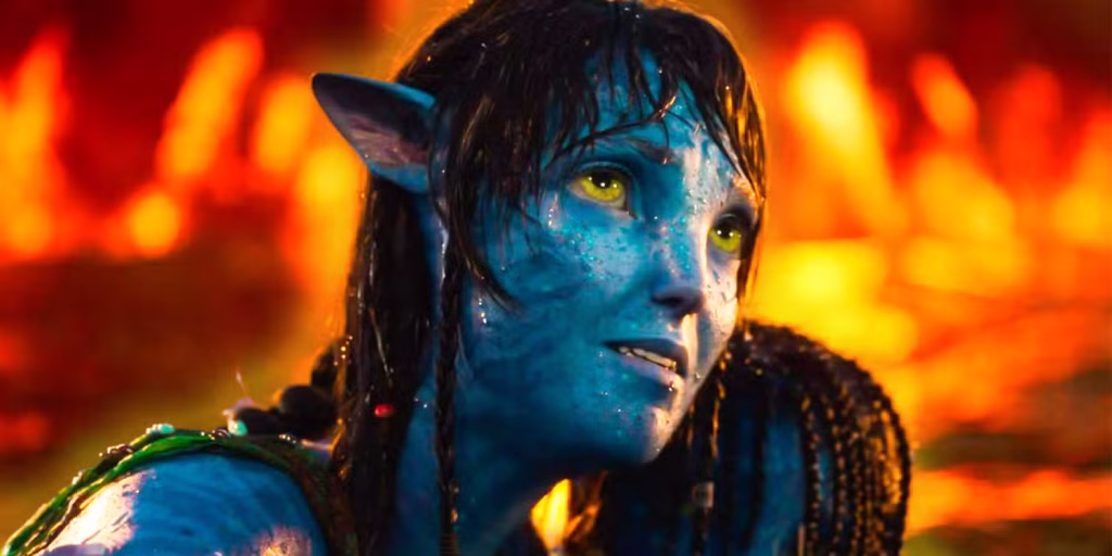 Close-up of a wet, blue-skinned Na’vi looking upward with bright flames blazing out of focus behind her.