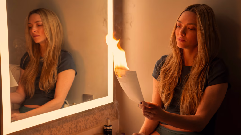 A blonde woman sits beside a lit vanity mirror, holding a sheet of paper that is burning at the top.