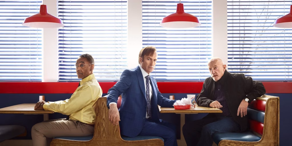 Gus Fring sits turned sideways in a diner booth while Jimmy McGill in a blue suit sits between booths and Mike Ehrmantraut leans forward, all three looking serious beneath red hanging lamps.