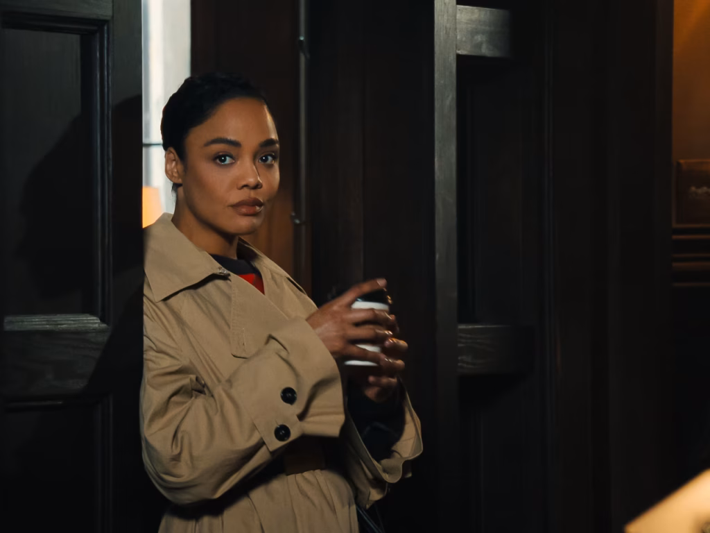 A woman in a beige trench coat stands in a doorway holding a drink and looks toward the camera in a dimly lit room.