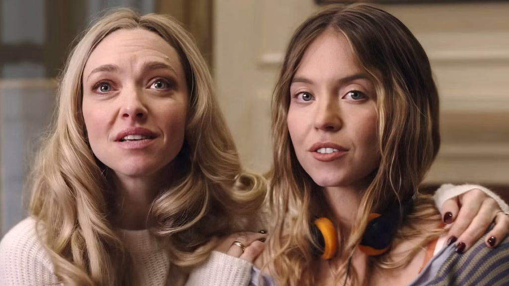 Sydney Sweeney and Amanda Seyfried sit close together indoors, with Sweeney wearing headphones around her neck while Seyfried rests an arm on her shoulder and looks on with a tense expression.