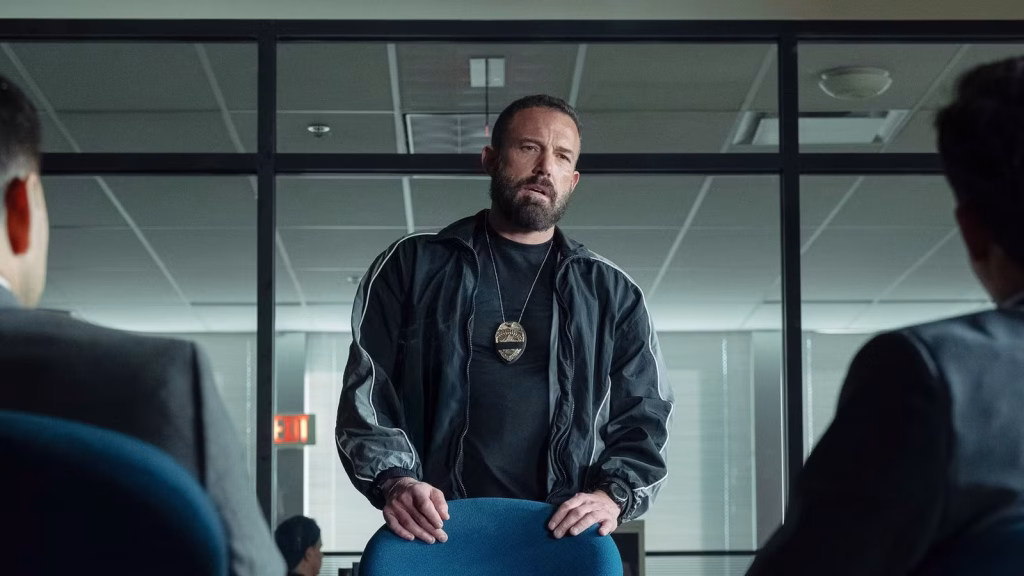 A bearded man wearing a police badge necklace stands behind a chair in a fluorescent-lit office, facing two people seated in the foreground.