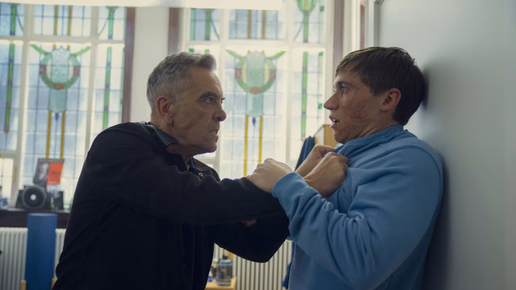 A man angrily grabs another man by the front of his hoodie and pins him against a wall while they stare at each other.