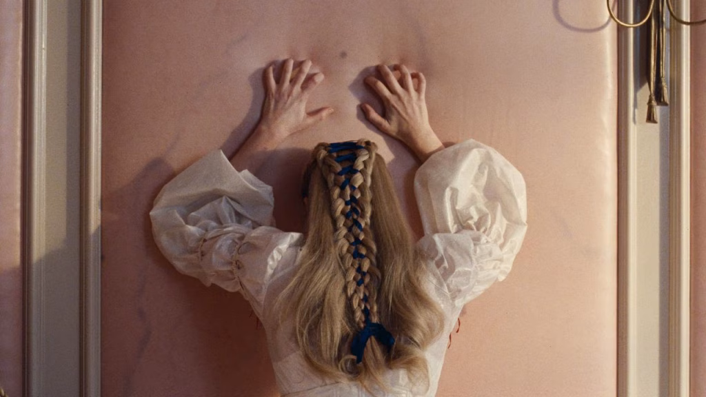 Rear view of Catherine Earnshaw with a long braided hairstyle pressing both hands against a pale pink wall during a tense scene in Wuthering Heights (2026).