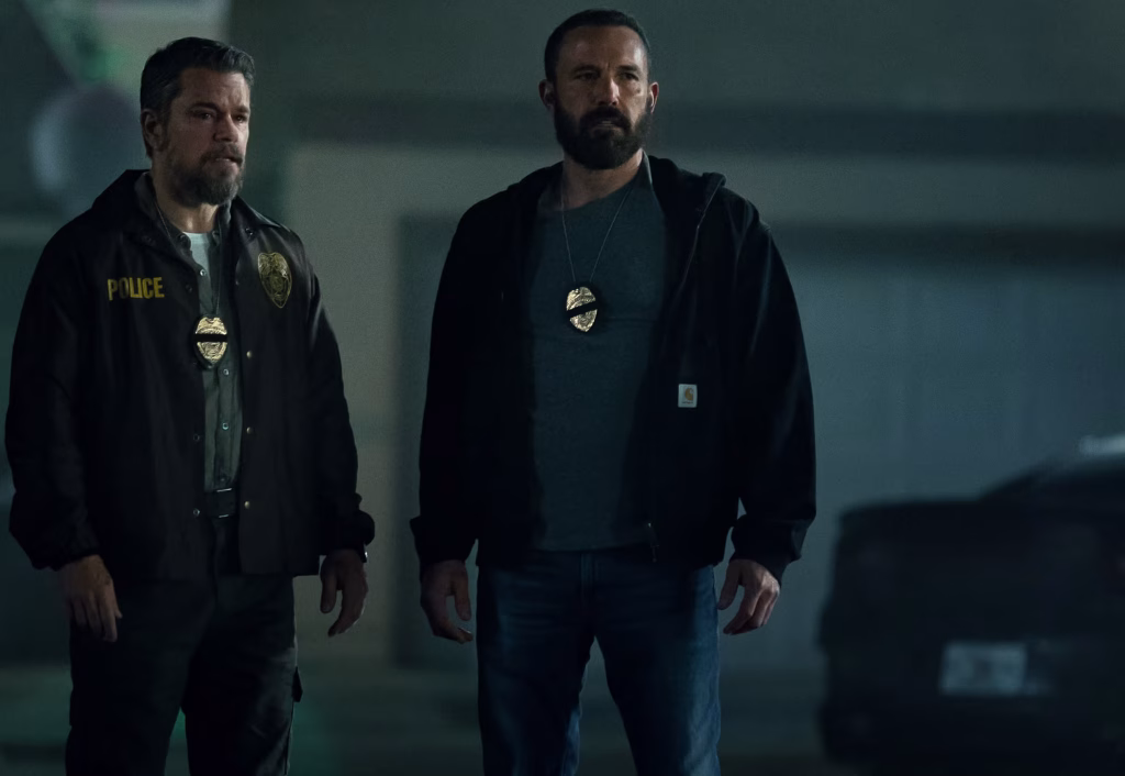 Two plainclothes detectives wearing police badges stand in a dim, hazy interior, staring ahead with tense, guarded expressions.