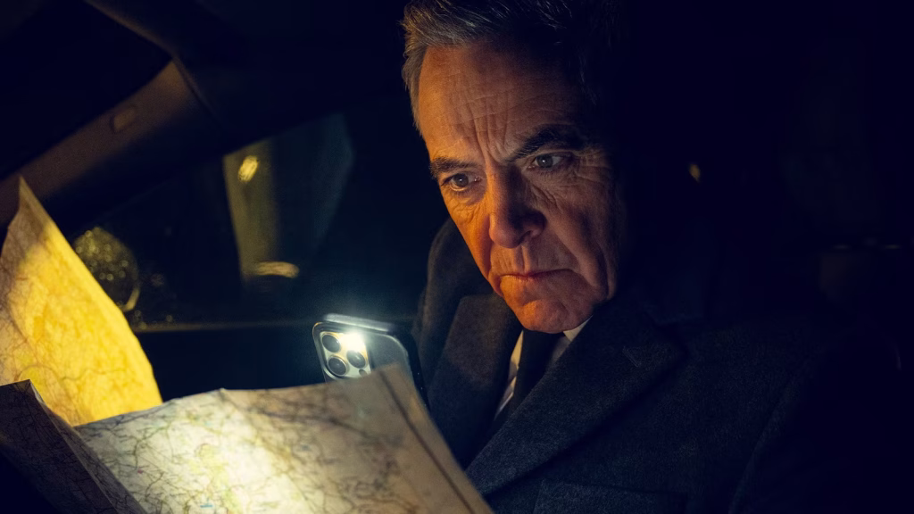 A man sits in a dark car at night, using his phone’s light to read a paper map with a tense, focused expression.