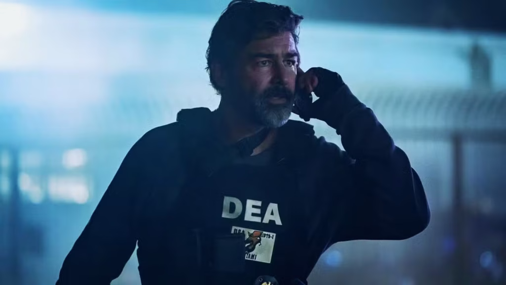 A bearded man in a black “DEA” hoodie holds an earpiece to his ear, looking alert in a blue-lit setting.