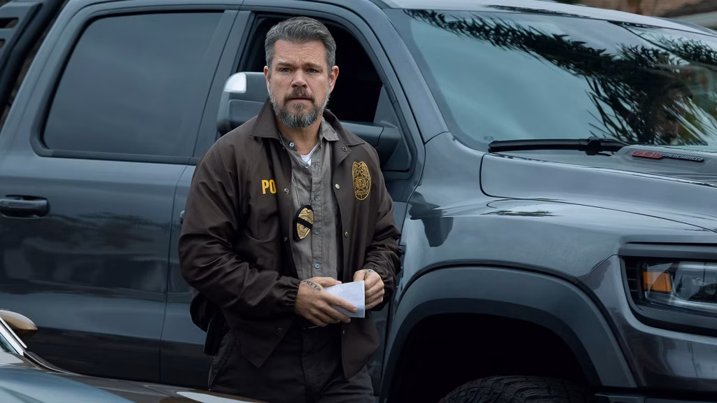 A police officer stands beside a dark pickup truck holding papers, wearing a badge and a brown jacket, and looks alert as if responding to an active scene.