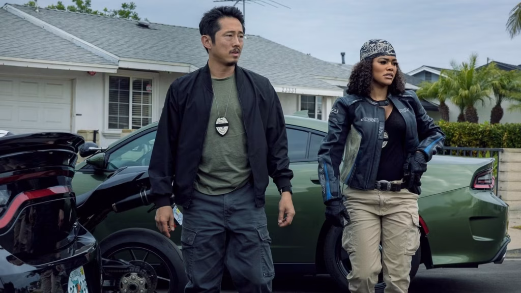 Two detectives wearing badges stand beside parked cars on a suburban street, looking off-camera with serious expressions.