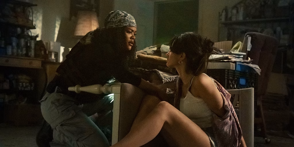 A detective crouches close to a woman sitting on the floor in a cluttered, dimly lit room, as they stare tensely at each other.