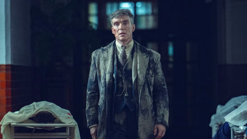 Tommy Shelby stands in a dim room wearing a dirt-covered suit, flanked by covered bodies on hospital trolleys in a grim scene from Peaky Blinders: The Immortal Man.