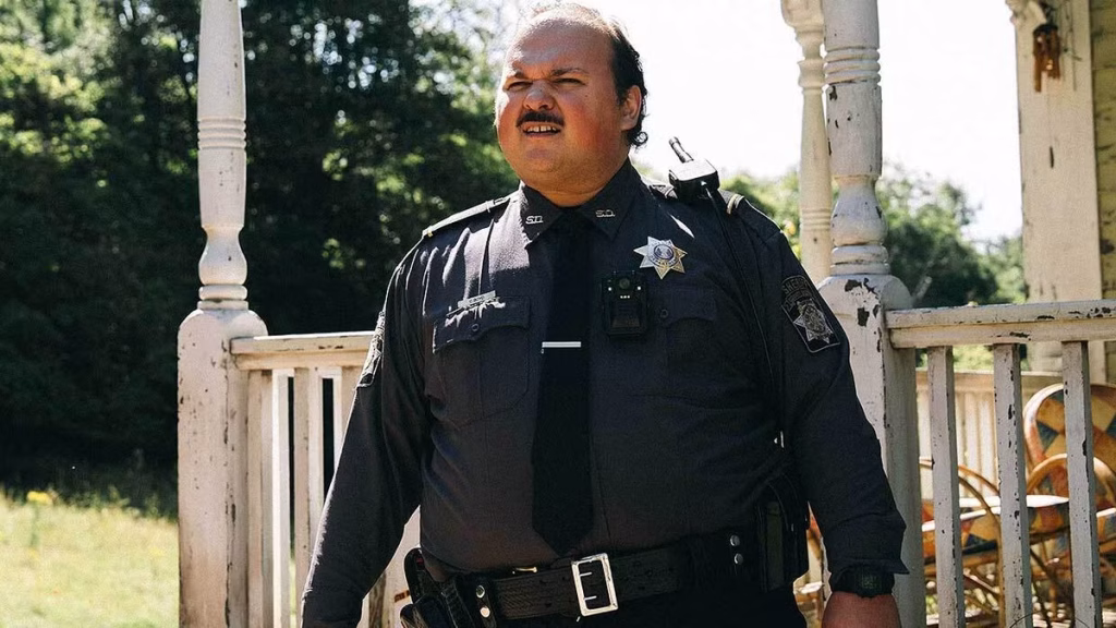 A uniformed officer stands outside on a sunlit porch in a scene from Bugonia.