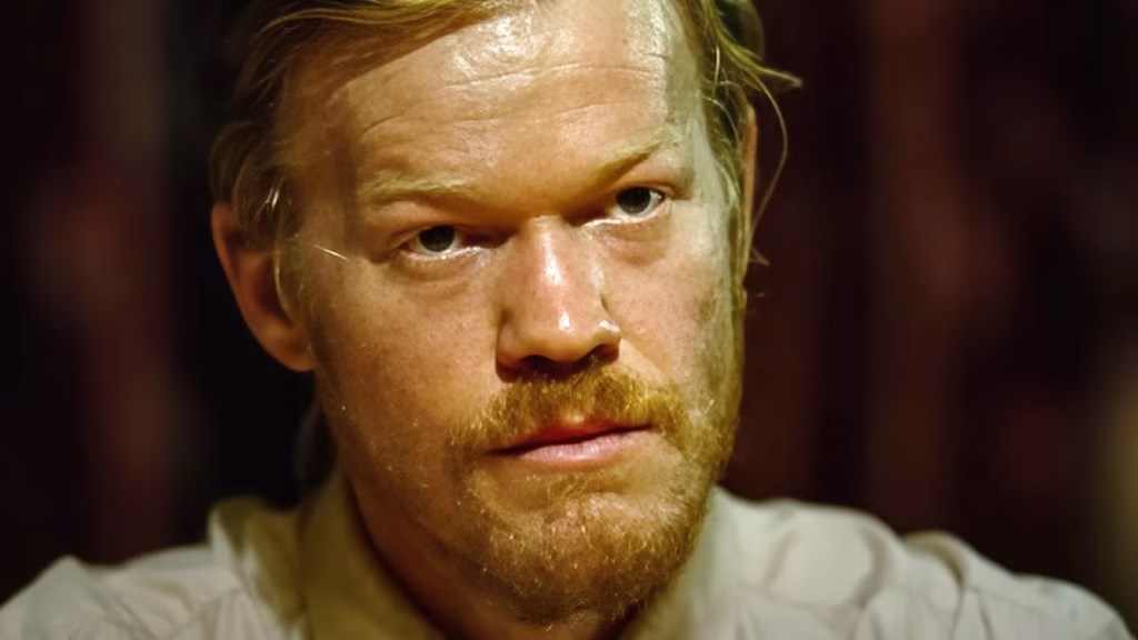 A close-up of Teddy in Bugonia shows him staring intensely ahead with a serious expression in a dimly lit setting.