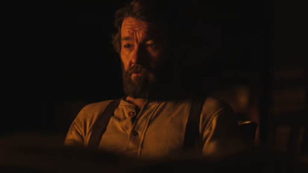 Robert Grainier sits indoors in a close-up scene from Train Dreams, looking thoughtful in work clothes.