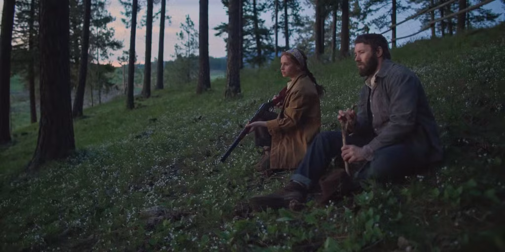 Gladys and Robert sit together on a grassy hillside in the woods in Train Dreams, with Gladys holding a rifle as they look out into the distance.