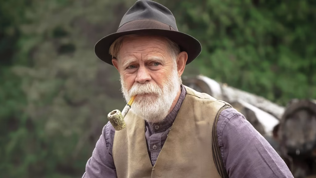 William H. Macy stands outdoors in period clothing and a hat, holding a pipe and looking off to the side in a thoughtful moment from Train Dreams.