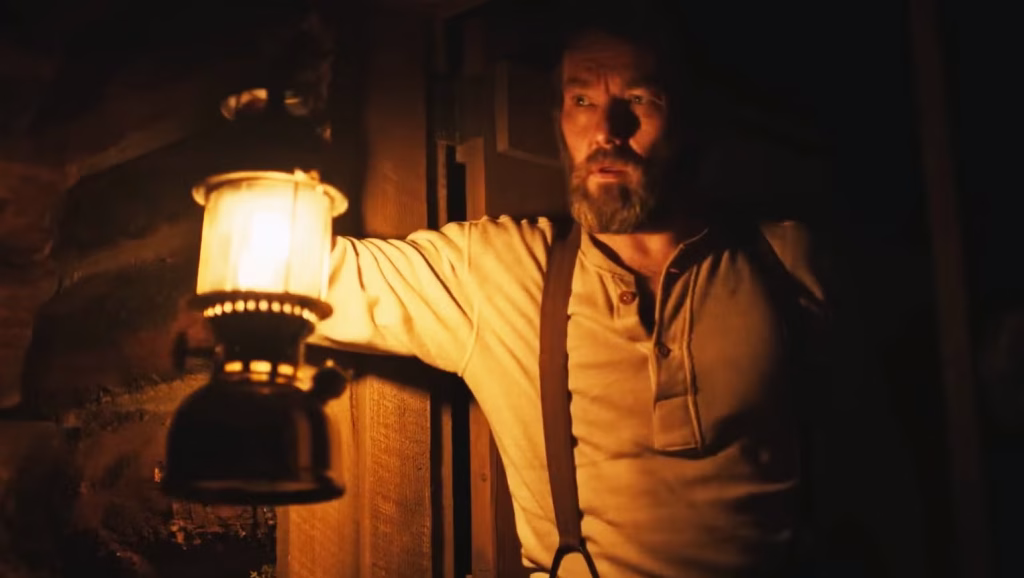 Robert Grainier stands in a dark doorway holding a glowing lantern in a nighttime scene from Train Dreams.