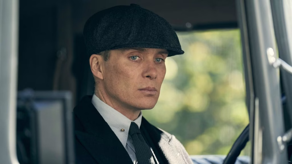 Tommy Shelby sits inside a car wearing a dark coat and flat cap, looking serious as he stares ahead.