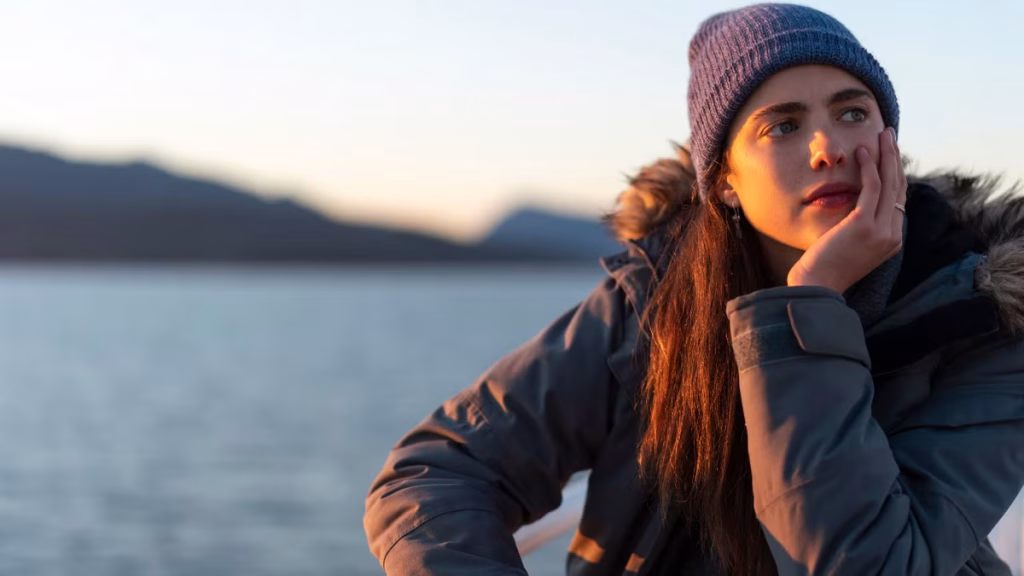 Margaret Qualley in a beanie and jacket, sitting in front of some water in Maid.