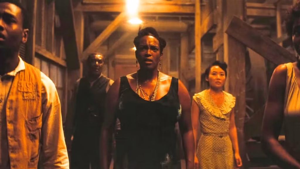 Several characters stand inside a dimly lit wooden building in a tense scene from Sinners.
