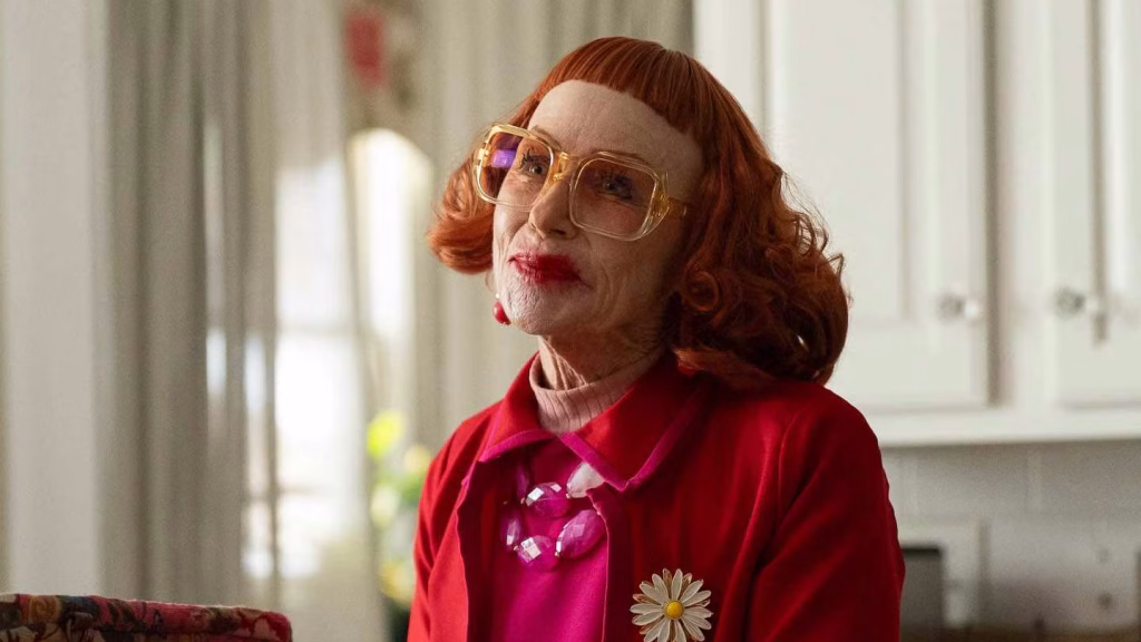Aunt Gladys sits indoors wearing red and pink with oversized glasses in Weapons.