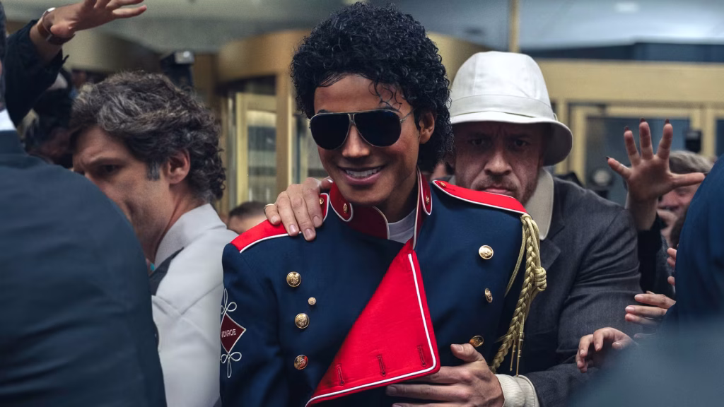 Jaafar Jackson as Michael Jackson smiling in a crowd while wearing sunglasses and a military-style jacket.