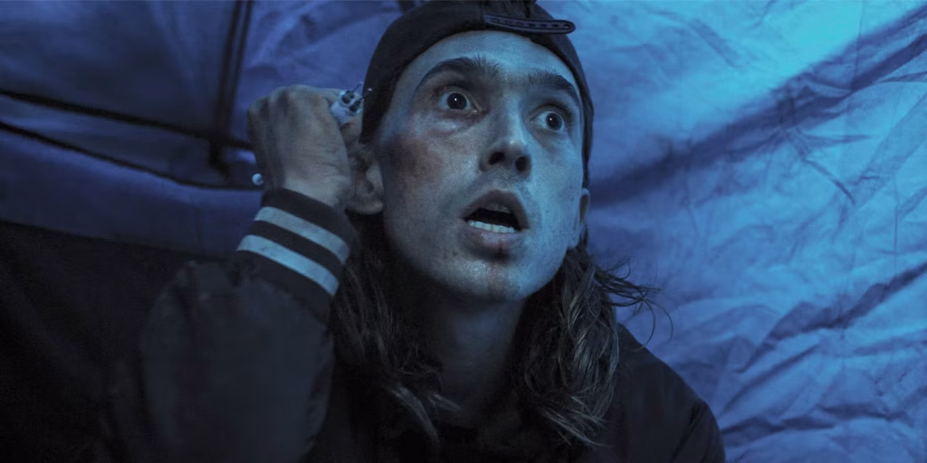 A frightened man in a backward cap looks upward under blue light in Weapons.