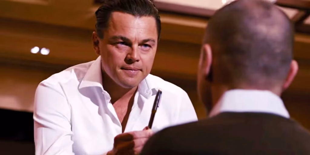Leonardo DiCaprio as Jordan Belfort holds a pen while speaking to another man.