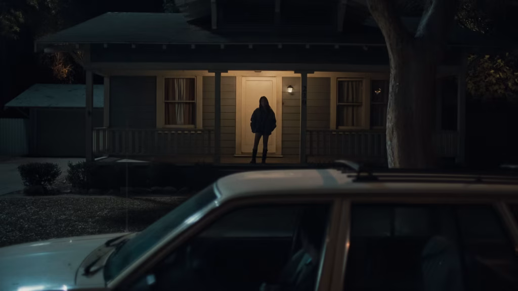 Nikki stands beneath a porch light outside Bear’s house at night in Obsession.