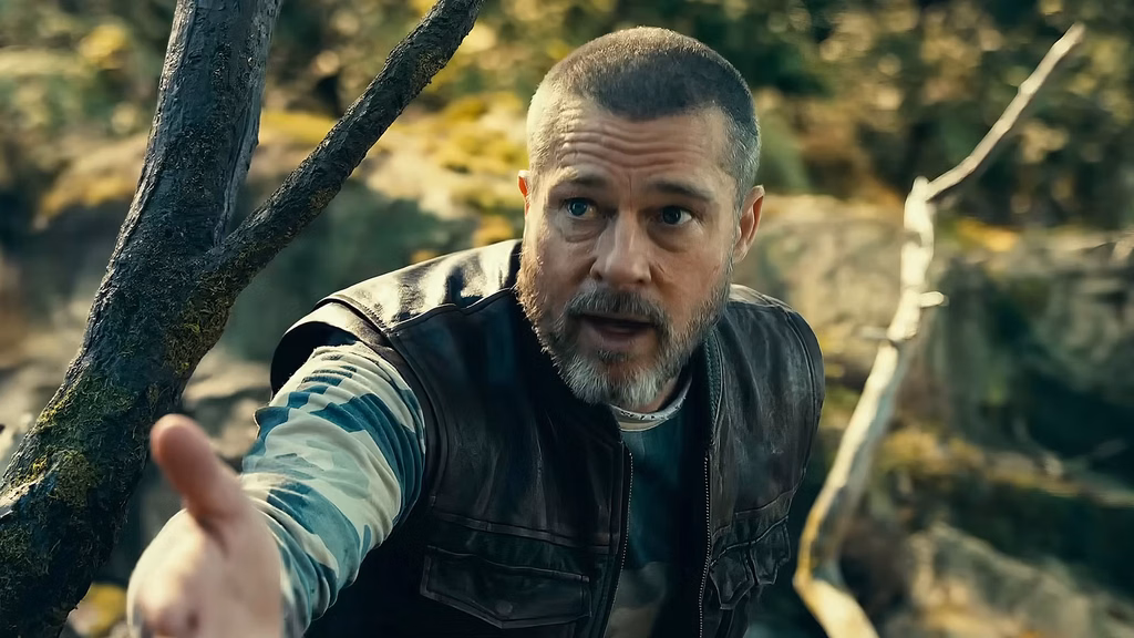 A bearded man in a dark vest and camouflage shirt extends his hand among tree branches in a wooded area.